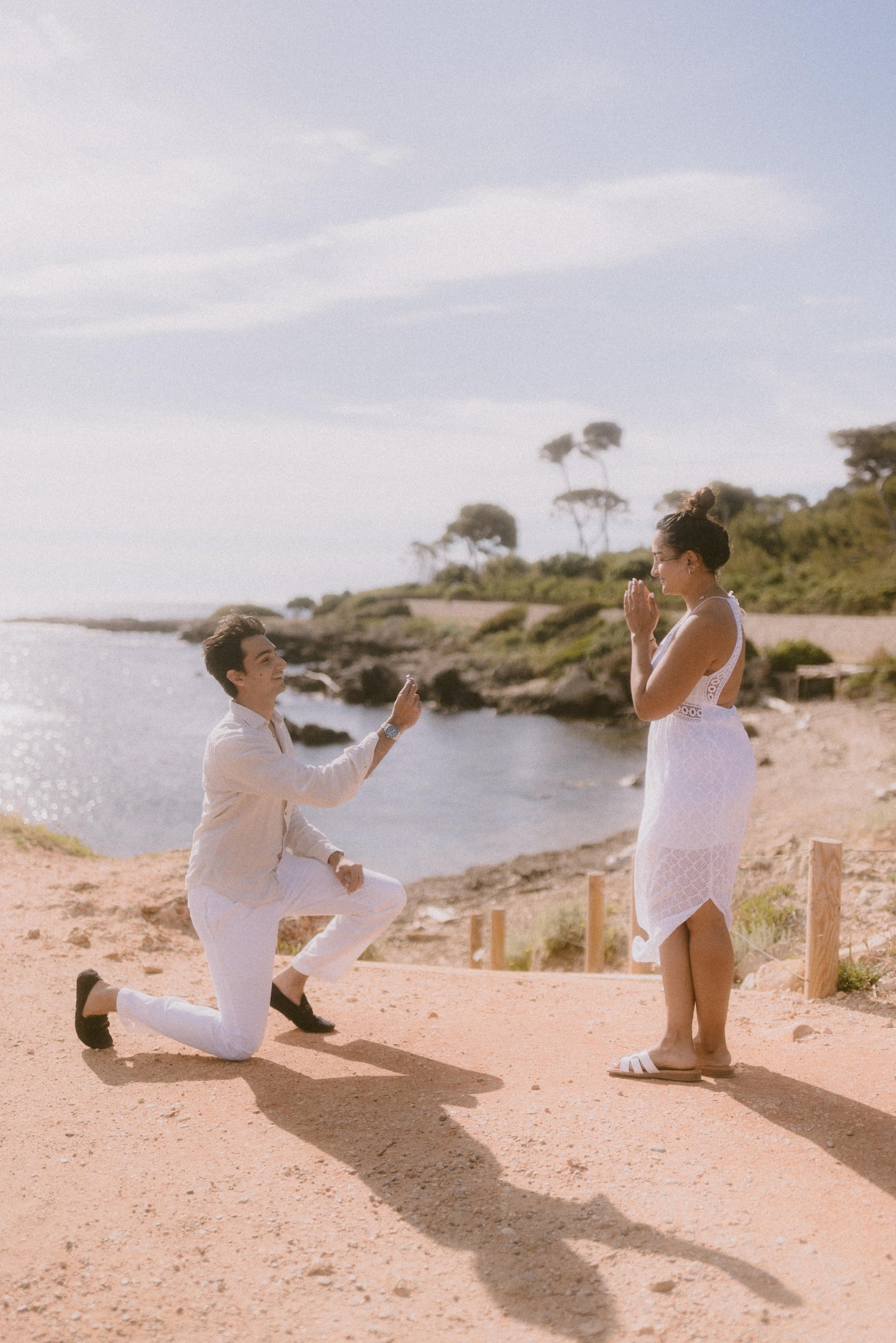 Proposal photographed by Terence Baelen, proposal and engagement photographer, at Cap d'Antibes, French Riviera. A man kneels down to propose to his girlfriend. There's a beautiful and scenic backdrop with a view over the sea. The girl is surprised.